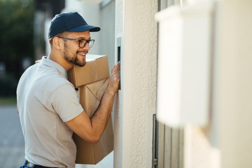 young happy deliverer with packages ringing intercom gate house